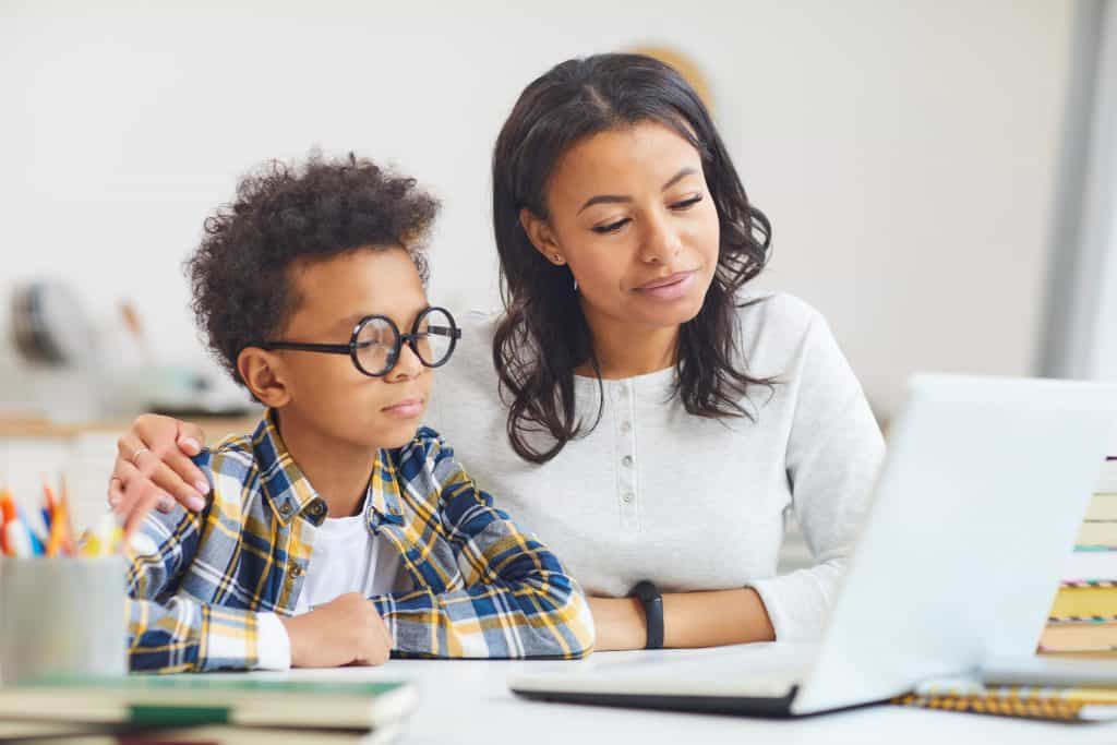boy wearing glasses while using laptop with mom, homeschooling and remote education concept